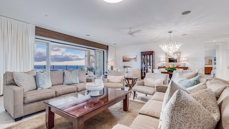 A bright, coastal living room with beige sofas, a wooden coffee table, large windows showing the ocean, and a chandelier in a stylish, open-plan layout.