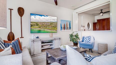 A bright coastal living room with a wall-mounted TV, white wood furniture, blue accents, and tropical decor in a cozy open-plan space.