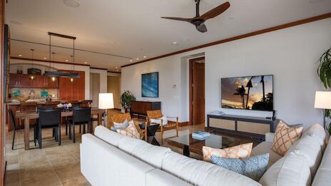 A bright, modern open-plan living room with a white sectional sofa, coffee table, and wall-mounted TV; adjacent kitchen/dining area, plants, and a ceiling fan, all in warm wood tones.
