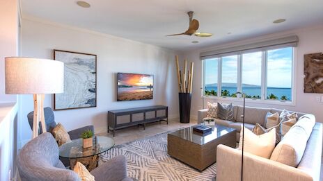 A bright, modern living room with a large window view, white walls, beige sofa, gray chairs, coffee table, wall art, and a ceiling bird statue.