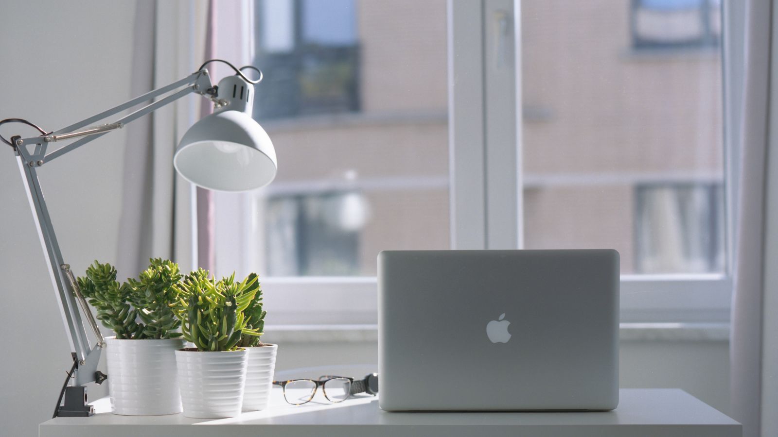 A modern workspace with a laptop, desk lamp, glasses, and potted plants on a white desk, positioned in front of a window.
