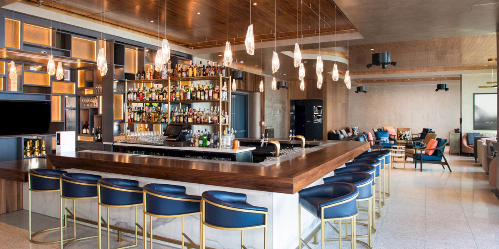 A modern bar with blue and gold stools, a well-stocked counter, hanging lights, and lounge seating in the background.