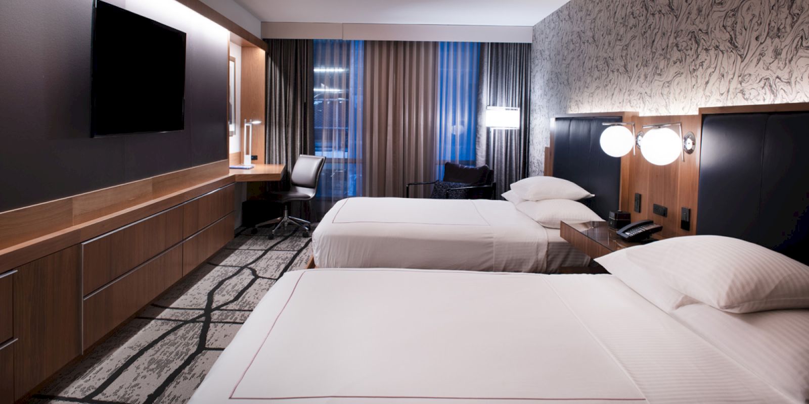 The image shows a modern hotel room with two double beds, a wall-mounted TV, a desk with a chair, and large windows with curtains, softly lit.