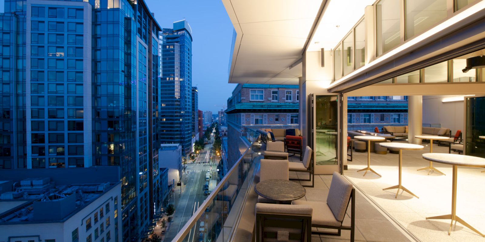 The image shows a modern cityscape at dusk with tall buildings and a stylish outdoor patio area featuring tables and chairs on a balcony.