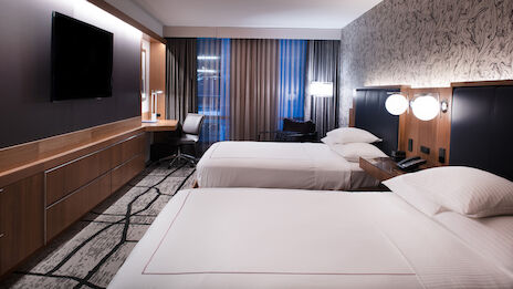 The image shows a modern hotel room with two neatly made beds, a desk and chair, a large TV, and stylish decor.
