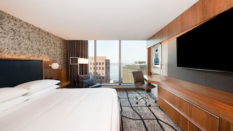 A modern hotel room with a large bed, wall-mounted TV, desk, chair, and a window offering a city view. The decor is sleek with wooden accents.