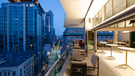 A modern balcony overlooking a city street at dusk, with outdoor seating and tables. The view includes tall buildings with illuminated windows.