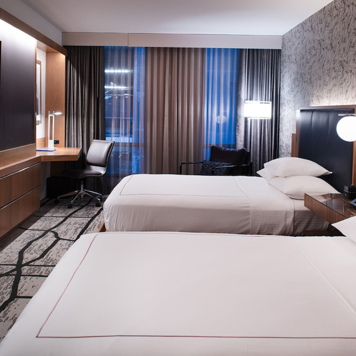 This image shows a modern hotel room with two double beds, a flat-screen TV on the wall, a desk with a chair, and minimalistic decor.
