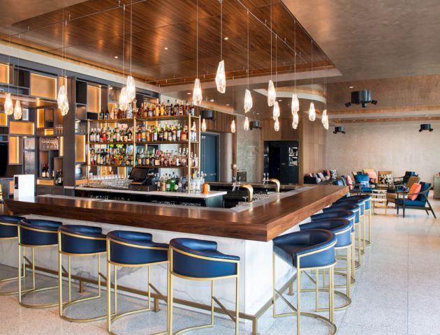This image shows a modern, well-lit bar with blue bar stools, a stocked liquor shelf, pendant lighting, and a seating area in the background.