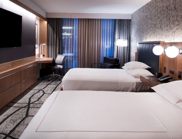 A modern hotel room with two beds, a wall-mounted TV, a desk with a chair, and stylish decor, illuminated by soft lighting and natural light from the window.
