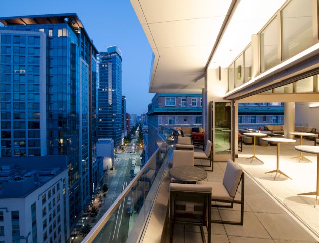 A modern balcony overlooking a cityscape during dusk with buildings, street views, and minimal outdoor furniture including tables and chairs.