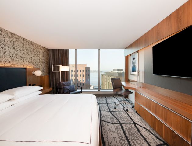 A modern hotel room with a large bed, a desk, a flat-screen TV, a chair, and a window with a city view. The decor is sleek and contemporary.