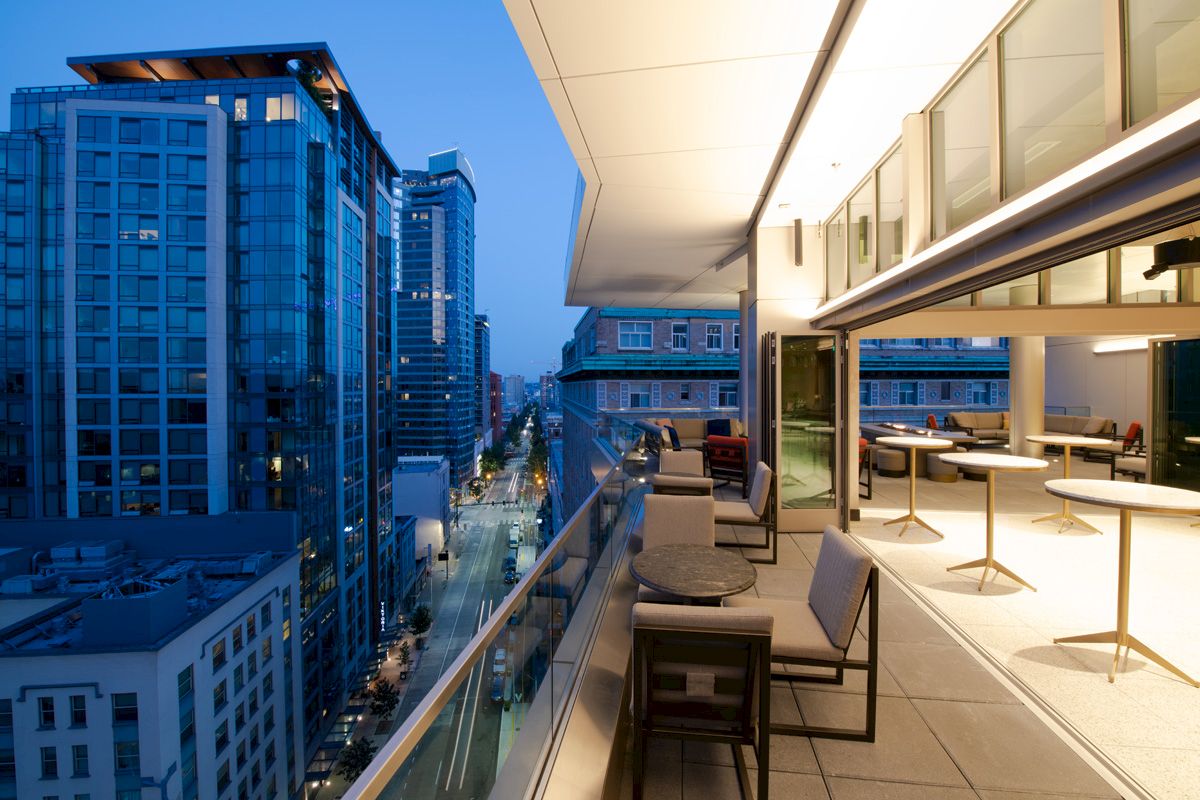 This image shows a modern urban balcony overlooking a cityscape at dusk, featuring seating areas and tables along the railing.