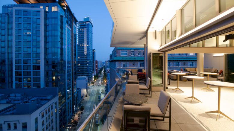 This image shows a modern urban balcony overlooking a cityscape at dusk, featuring seating areas and tables along the railing.
