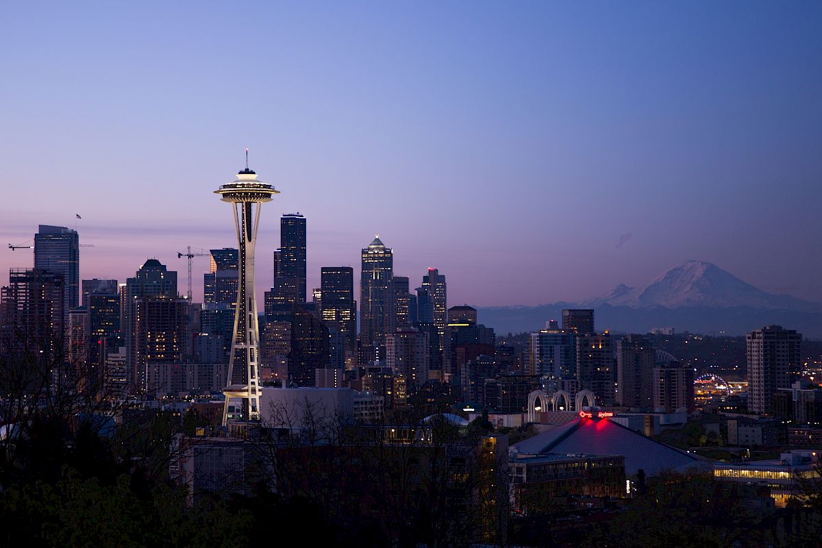 The image shows the Seattle skyline at dusk, featuring the Space Needle prominently, with Mount Rainier visible in the background and city lights illuminating.