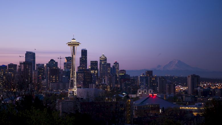The image shows the Seattle skyline at dusk, featuring the Space Needle prominently, with Mount Rainier visible in the background and city lights illuminating.