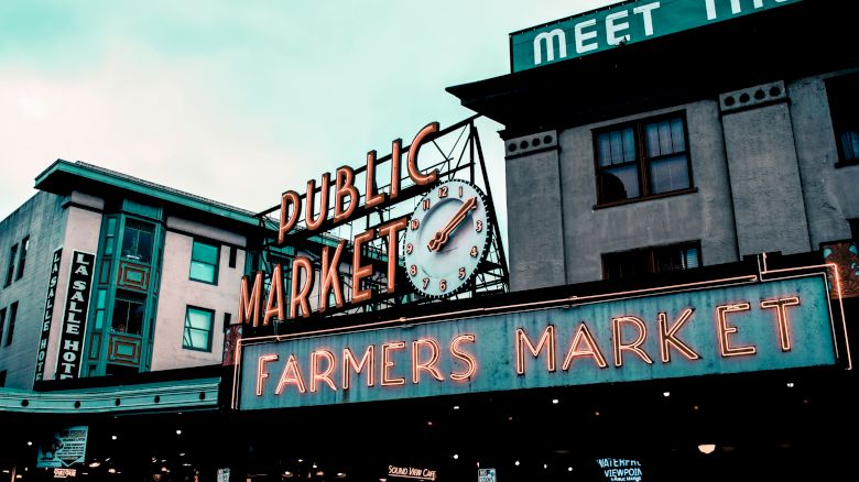 The image shows a neon sign for a Public Farmers Market with an old-fashioned clock. Buildings in the background have additional signage.