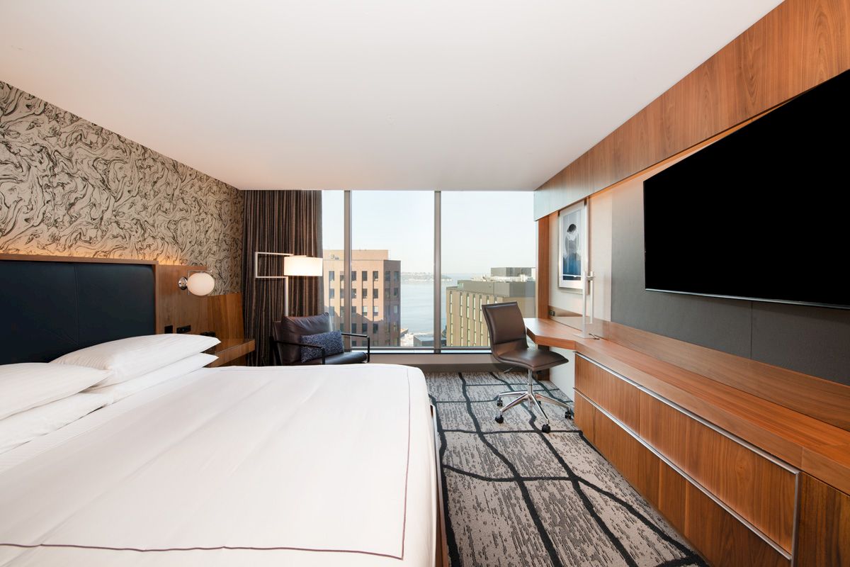 The image shows a modern hotel room with a large bed, TV, desk, chair, city view window, and contemporary decor.