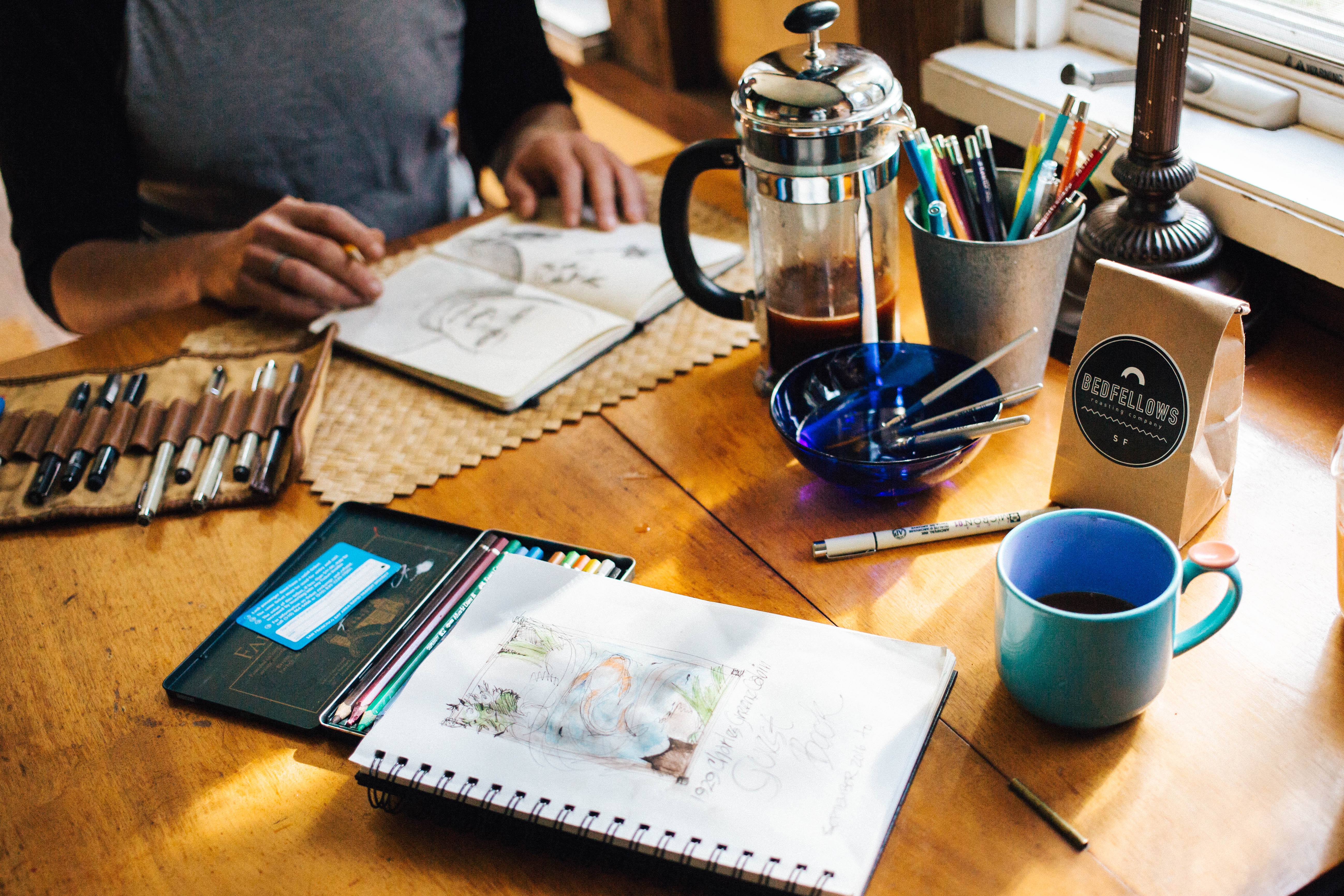 A person is sketching at a wooden table with drawing materials, a coffee French press, a cup of coffee, and a container holding various pencils.